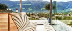 Wooden bench in a room with panoramic glass windows and views of a village below, and surrounding mountain peaks
