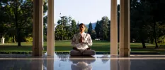 Woman in white clothes in lotus position with her hands clasped in prayer in a columned pavilion with a field behind