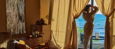 Woman stands in a robe looking out to sea from her terrace windows, with ivory drapes cascading from the walls and wooden furniture inside