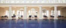 White-robed woman walks past an indoor pool surrounded by white pillars and rattan loungers with navy cushions