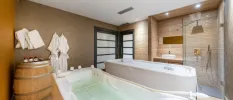 Spa treatment room with a shower, bath and massage bed, a wooden barrel with bottles and white towels and robes hung on the wall