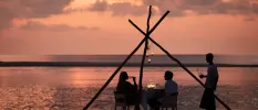 Silhouette of a couple sipping wine at a table by the shoreline as the sun sets and a staff member stands nearby