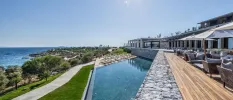 Outdoor pool and wooden terrace with tables, chairs and parasols, overlooking landscapes grounds and the Aegean Sea