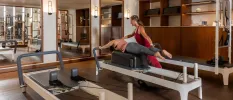 Woman lies on her front on a reformer Pilates machine as a staff member in red active wear supports her posture