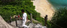 Staff member walks down a curved hillside staircase carrying a tray, as the waves lap a golden sandy beach below