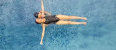 Woman in a black swimsuit and swim hat floats on her back in a pool, supported by another woman