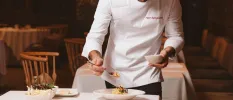 Man in chef whites spooning food onto a white plate, on a table with a white tablecloth