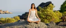 Woman sitting by the ocean practising meditation on rocks