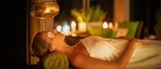 Woman relaxing in a towel on a massage bed with eye pads on and candles surrounding her