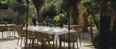 Table with white table cloth and wooden seats laid out for a meal in a lush green courtyard