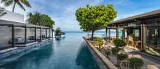 Infinity pool with ocean views and trees, lined on one side by grey-cushioned daybeds and the other by a leafy terrace with rattan chairs and tables