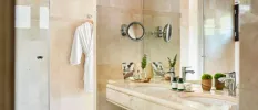 Cream marble bathroom with a white robe hanging on the wall, a vanity unit with two sinks and potted green plants, a shower and a large mirror, plus a magnifying mirror coming out of the wall
