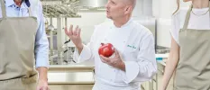 Chef in whites holding a red vegetable gesturing with two people in aprons next to him