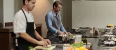 Two men in aprons stand at hobs while enjoying a cooking class, with fresh ingredients around them