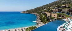 Low-rise villas, an infinity-edge pool and terrace with white-cushioned loungers and umbrellas, on a hillside overlooking a sandy beach furnished with loungers and umbrellas and overlooking a turquoise sea