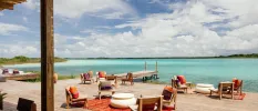 Wooden deck on the waterfront with wooden chairs with orange cushions, orange blankets and bean bags overlooking the lagoon