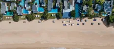 Aerial view of a sandy beach, white villas with private pools and surrounding greenery