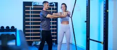 Woman in light leggings and a lilac gym top stretches a rope in a gym, supported by a personal trainer