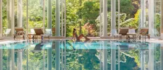 Toned woman in a swimsuit sits at the edge of an indoor pool surrounded by white pillars and wooden, white-cushioned loungers, with floor-to-ceiling glass in the background showcasing the gardens beyond