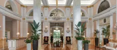 High ceilinged lobby area with ornate rugs and armchairs, gold accents, glittering chandelier-stye wall lights and grand white pillars