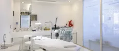 Medical-style treatment room with a machine with wires, unit filled with bottles, a bed covered in white towels and gleaming white walls and flooring