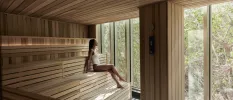 Woman in a white swimsuit sits in a wooden sauna looking out a full-length window at tropical greenery