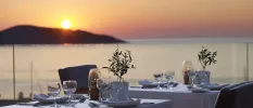 Elegant table set-up with crisp white tablecloths, glassware and olive cuttings on a sea-facing terrace with a glass balcony, as the sun dips behind a hillside opposite