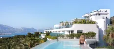 Blonde woman in a black bikini sits with her back to the camera on an infinity-edge pool overlooking lush greenery and mountains