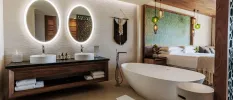 Bathroom with a large window overlooking the bedroom, with twin sinks, mirrors, a standalone bathtub, and dark-wood furnishings with a macrame design on the wall