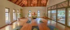 Airy studio with rattan lighting, a wood-beamed ceiling and floor-length windows set up with yoga mats and cushions ready for a class