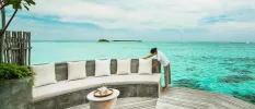 Staff member adjusts an ivory cushion on a cushioned stone sofa, on a wooden deck with direct access to the turquoise ocean