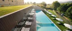 Long, rectangular swimming pool surrounded by white daybeds, loungers and umbrellas, next to grassy lawns and overlooking the countryside