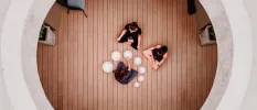 Two women in black sit cross legged in a wood-floored studio while a professional surrounded by bowls carries out sound healing