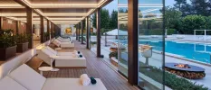 Wooden decking with white daybeds and glass windows overlooking firepits and a swimming pool