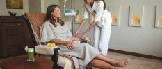 Woman in white sits with a drip in her arm smiling as a staff member assists her
