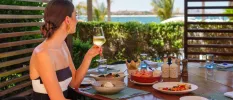 Woman in striped strapless top and gold earrings sips white wine with a sea view, and a table filled with seafood