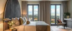 Hotel room with a stylish curved velvet headboard, white bedding with neutral cushions, floor-to-ceiling windows with neutral curtains and a desk area with a gold vase and flowers
