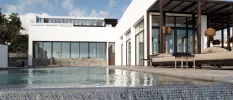 Infinity pool edge next to cushioned loungers overlooked by a building with full-length windows
