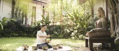 A woman sits on a blanket in a tropical garden, using gold sound healing bowls in front of a Buddha statue