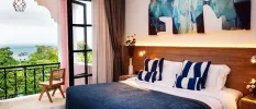 Hotel room with white bedding and walls, a navy throw and blue and white cushions, a wooden headboard and a blue and white wall painting, plus balcony doors open onto a balcony with greenery and sea views