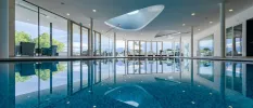 Large indoor pool surrounded by white-cushioned loungers under a white ceiling with spotlights, and floor-to-ceiling windows showing the greenery outside