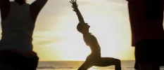 Silhouette of three people in yoga poses on the beach as the sun sits just above the ocean horizon