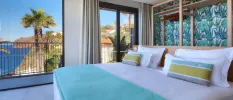 Modern hotel room with white bedding with a blue throw and striped cushions, tropical wallpaper and a view of the sea and cliffs