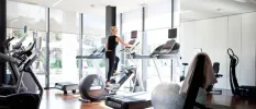 Woman in black works out on a machine in a gym with full-length windows, bikes, treadmills and an exercise ball
