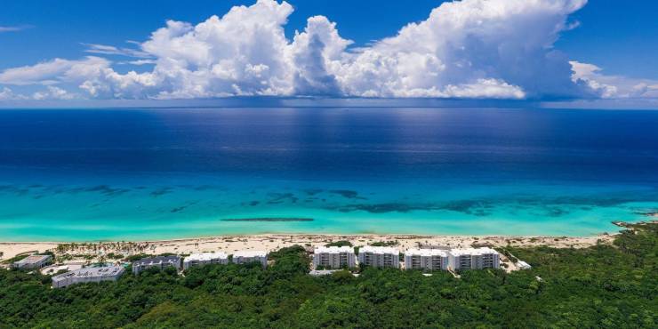 Aerial view of Palmaïa – The House of AïA Thick green forest next to a high-rise white resort, with white sands and turquoise shallows in front of it, under a blue and cloud sky