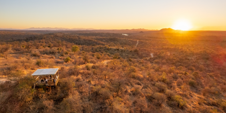 The sun sets over the South African wilderness while a group enjoys the view from a wooden deck