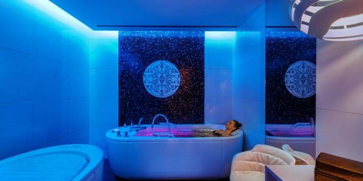 Woman in black swimwear relaxing in a filled bathtub in a white room with atmospheric blue mood lighting