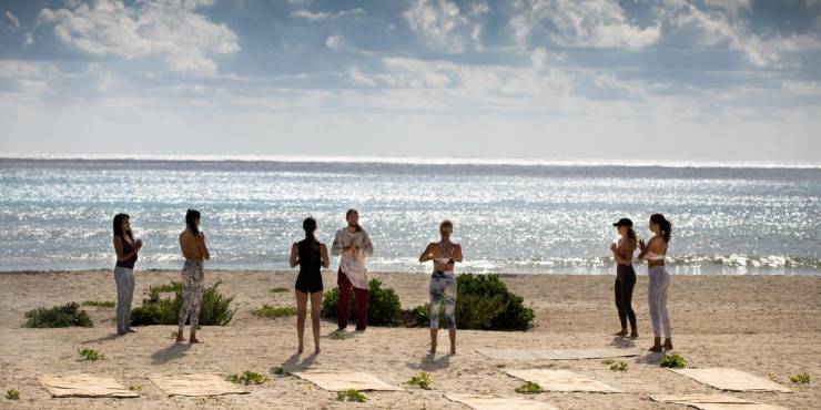 Group yoga at Palmaïa – The House of AïA Group of women in active wear stand on the beachfront in front of mats with their hands clasped in prayer, as a yoga instructor leads from the front
