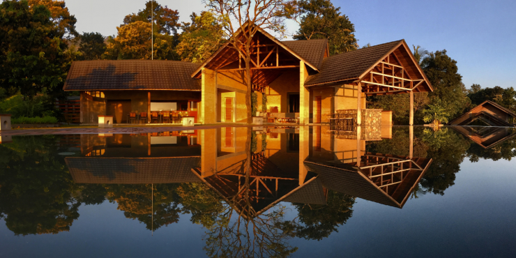 Lake-like swimming pool under a blue sky among tropical greenery, overlooked by a wooden building with triangular rooftops and stilts