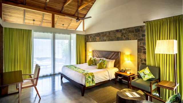 Hotel bedroom with a stone wall, green curtains, woof-beamed ceiling, white bedding with green soft furnishings, a seating and desk area, tiled floors and and floor-to-ceiling doors